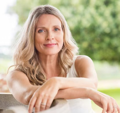 Cuidado Sênior – Mulher de meia-idade com cabelos loiros ondulados, sentada ao ar livre, com expressão serena e confiante.