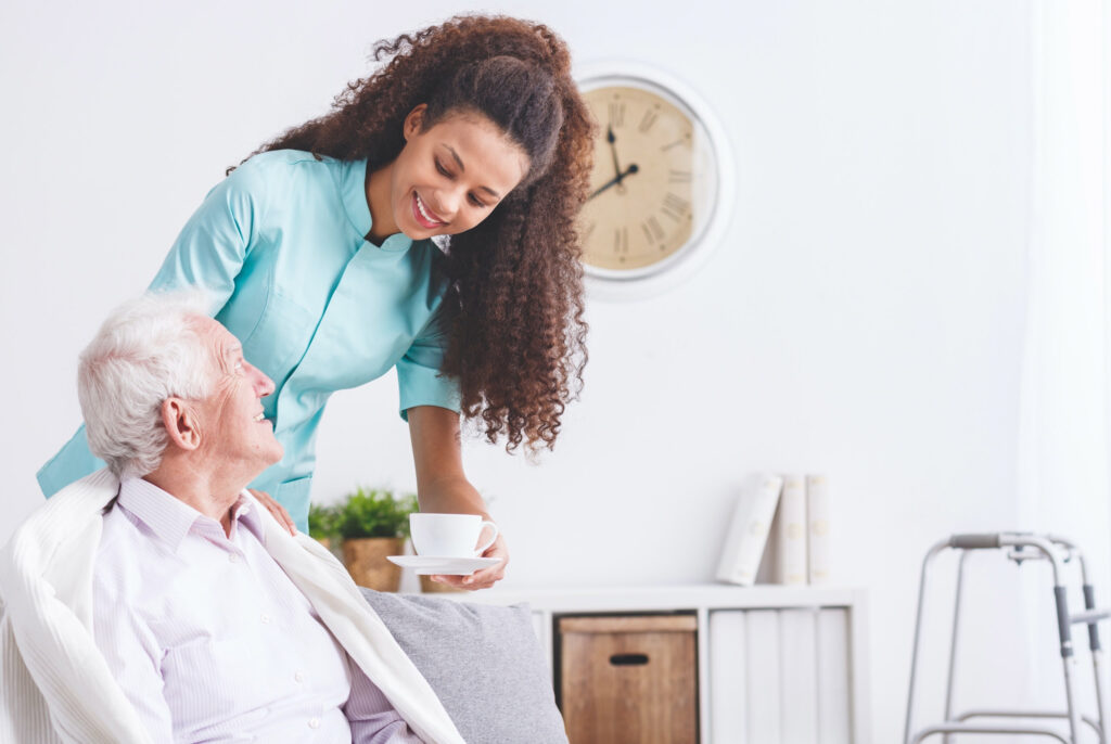 Cuidado Sênior – Cuidadora sorridente entrega uma xícara a idoso sentado, ambos trocando olhares afetuosos em ambiente claro e acolhedor.