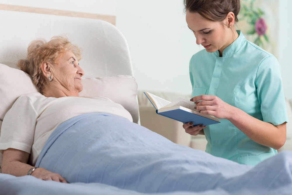 Cuidado Sênior – Cuidadora hospitalar lê para idosa acamada, proporcionando companhia, estímulo cognitivo e acolhimento em ambiente tranquilo.