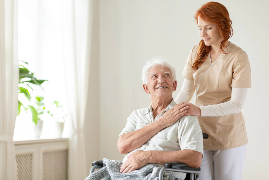 Cuidado Sênior - Cuidadora Residencial toca com carinho o ombro de idoso em cadeira de rodas, ambos sorrindo em ambiente claro e acolhedor.