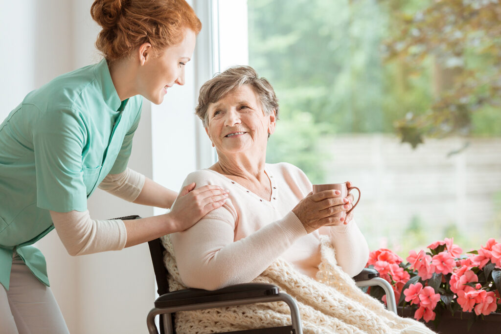 Cuidado Sênior - Cuidadora sorri para idosa em cadeira de rodas que segura uma xícara, coberta com manta, próxima a uma janela com flores.