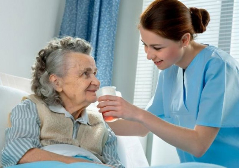 Cuidado Sênior - Cuidadora Residencial sorri ao oferecer copo com medicamento a idosa sentada na cama, em ambiente acolhedor e iluminado.