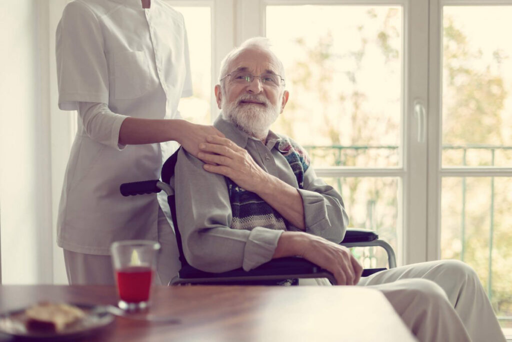 Cuidado Sênior - Idoso em cadeira de rodas é acolhido por cuidadora, com expressão serena e olhar de confiança, em ambiente residencial iluminado.