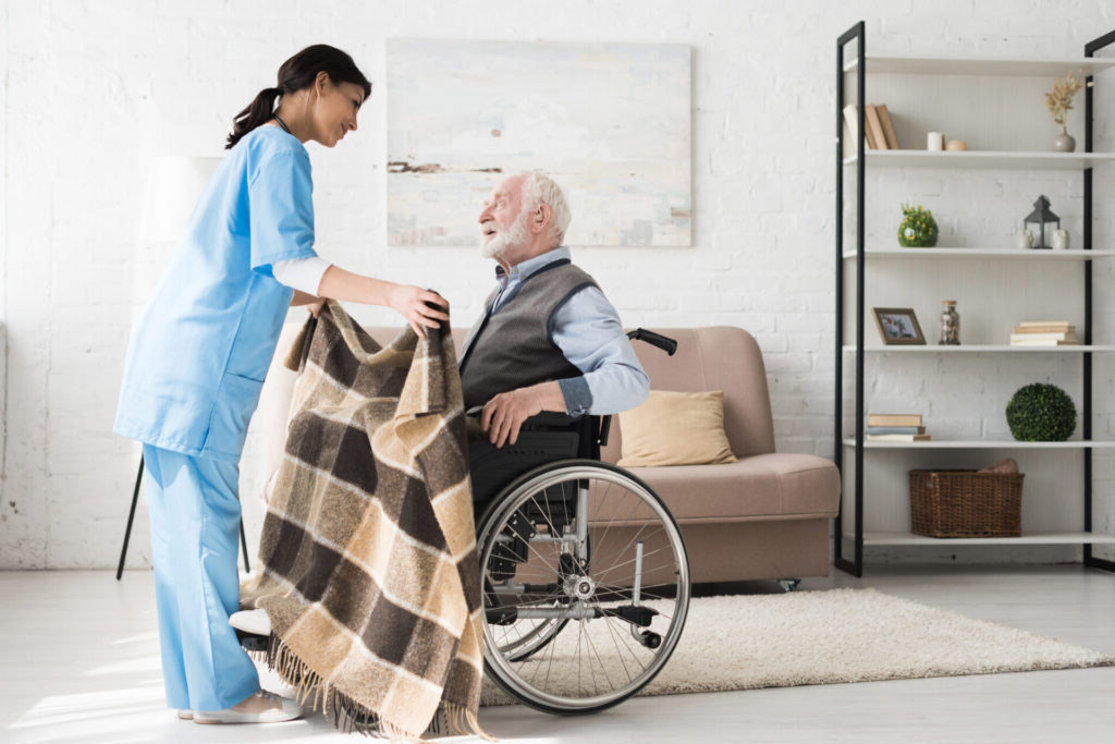 Cuidado Sênior - Cuidadora Residencial cobre idoso em cadeira de rodas com manta xadrez, em ambiente residencial aconchegante e iluminado.