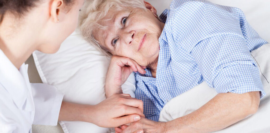 Cuidado Sênior - Idosa acamada olha com ternura para cuidadora, que segura sua mão com carinho em ambiente acolhedor.