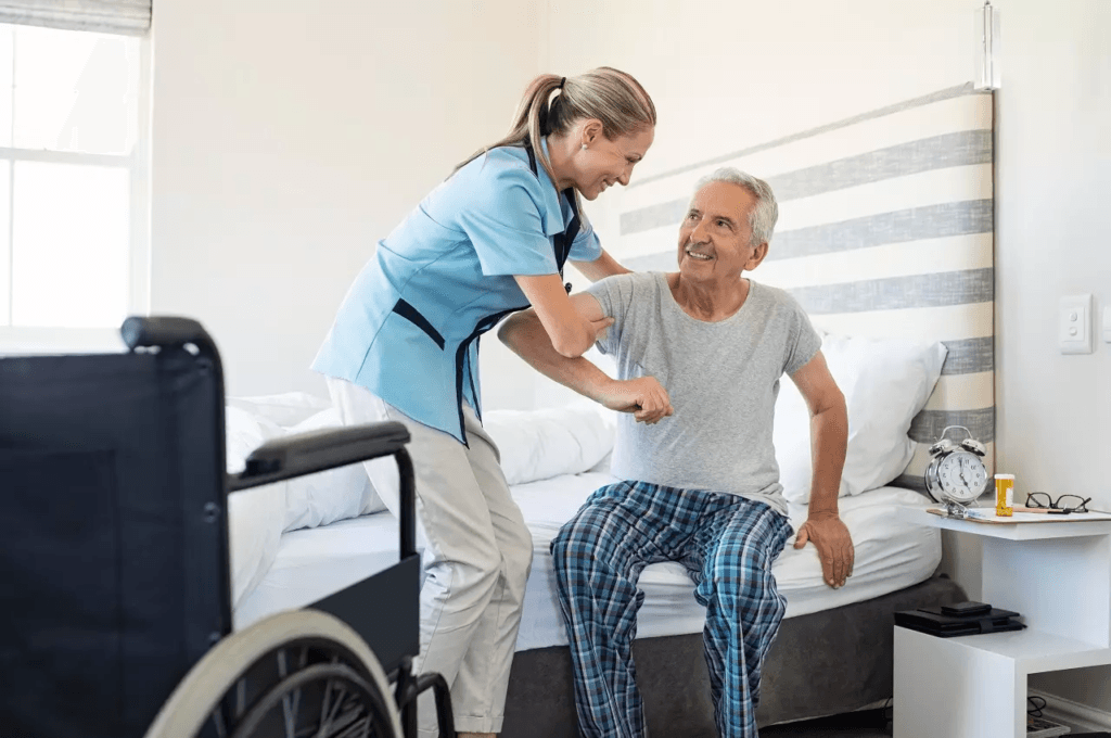 Cuidado Sênior - Cuidadora ajuda idoso a se levantar da cama com apoio e sorriso, ao lado de uma cadeira de rodas em ambiente claro e organizado.