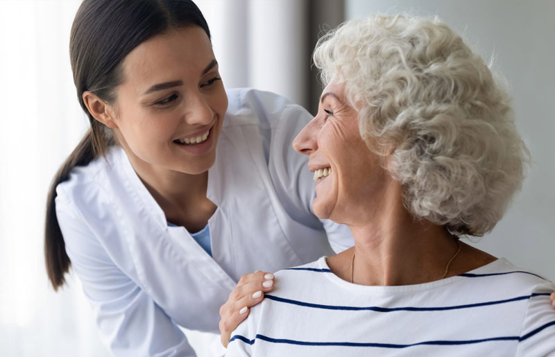 Cuidado Sênior - Cuidadora sorridente olha com carinho para idosa de cabelos grisalhos, ambas felizes e conectadas em ambiente iluminado.