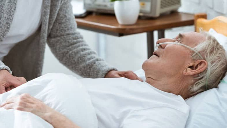 Cuidado Sênior - Cuidadora hospitalar ao lado de idoso acamado com cateter nasal, oferecendo atenção em ambiente calmo e confortável.