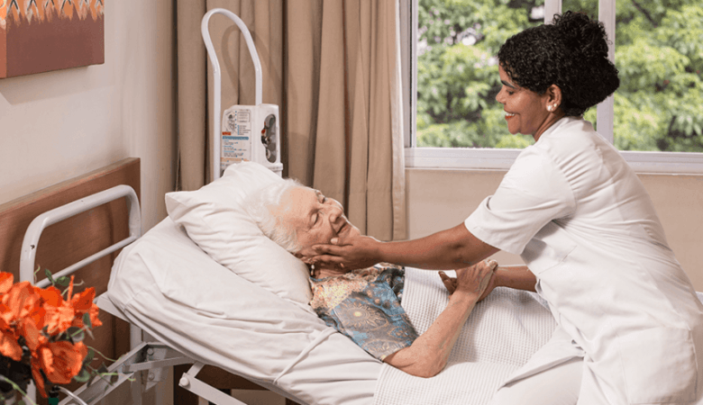 Cuidado Sênior - Cuidadora hospitalar acaricia o rosto de idosa acamada, demonstrando afeto e atenção em quarto iluminado com vista para a natureza.